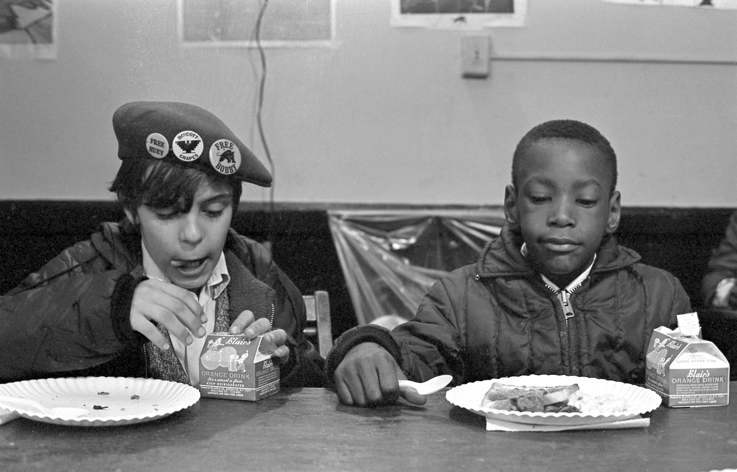 A Black boy and White boy sit side-by-side eating breakfast.
