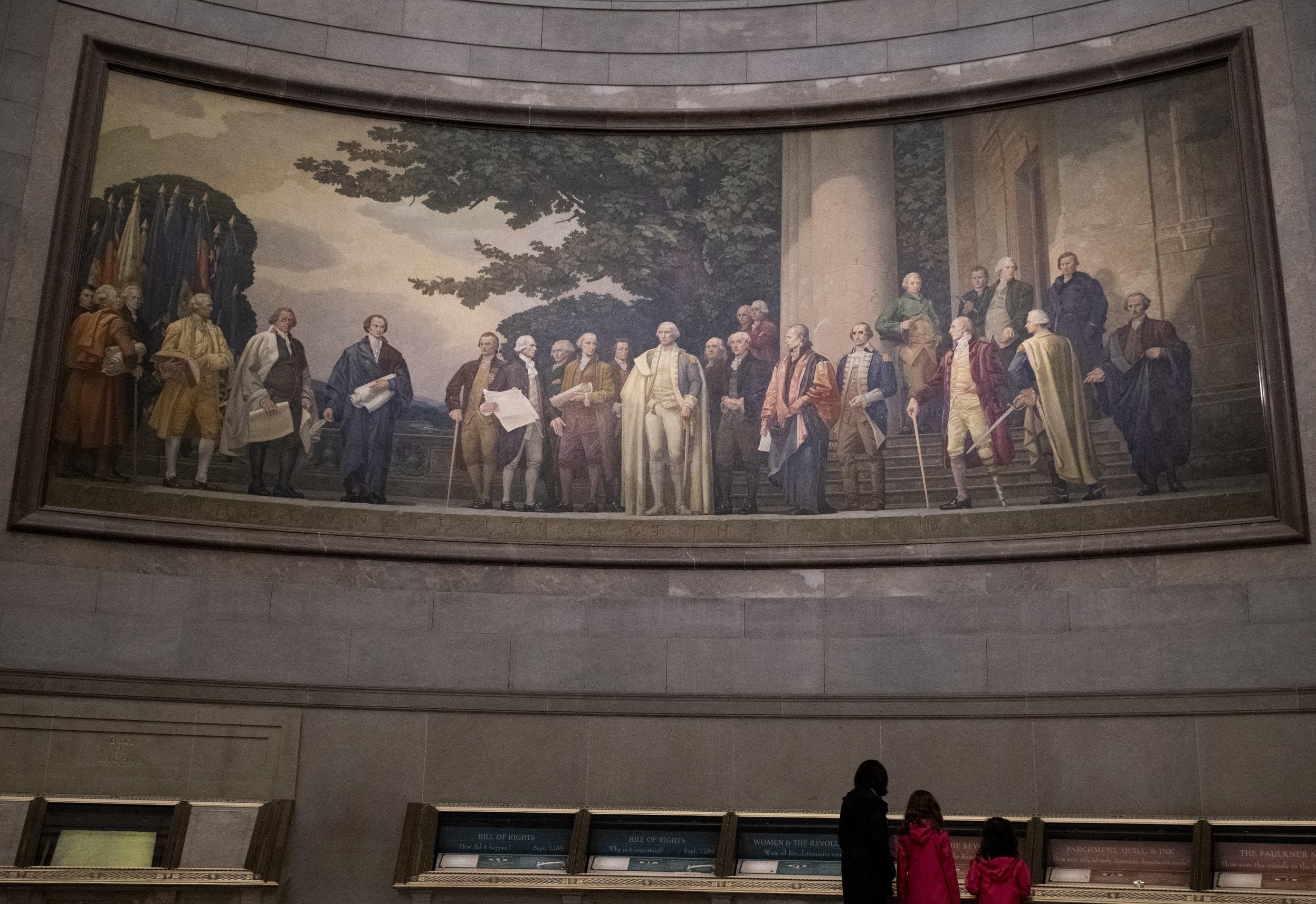 A large mural inside the National Archives Museum shows the delegates standing outdoors by a building with large columns. George Washington appears in the middle with the men's attention focused on him. A few of the delegates hold large documents in hand.