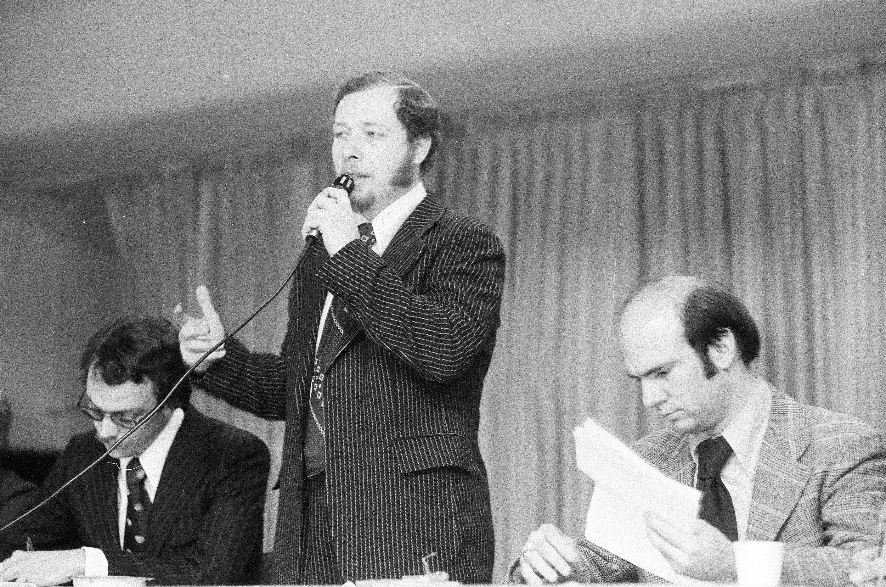 A black and white photo shows Cha Cha Jiménez standing at a table and speaking through a microphone. Two White men sit on either side looking down at their papers.