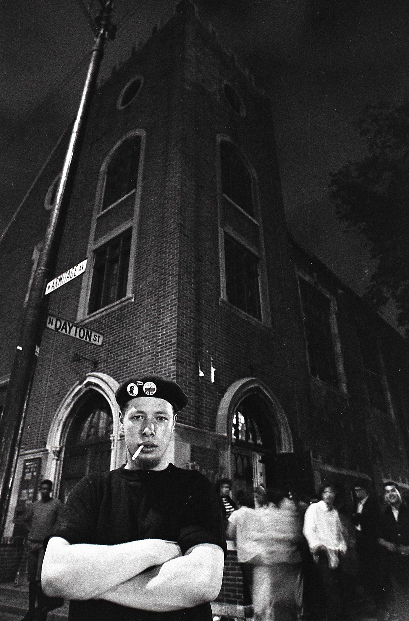 A black and white photo of José “Cha-Cha” Jiménez standing with arms crossed in front of a church at night.