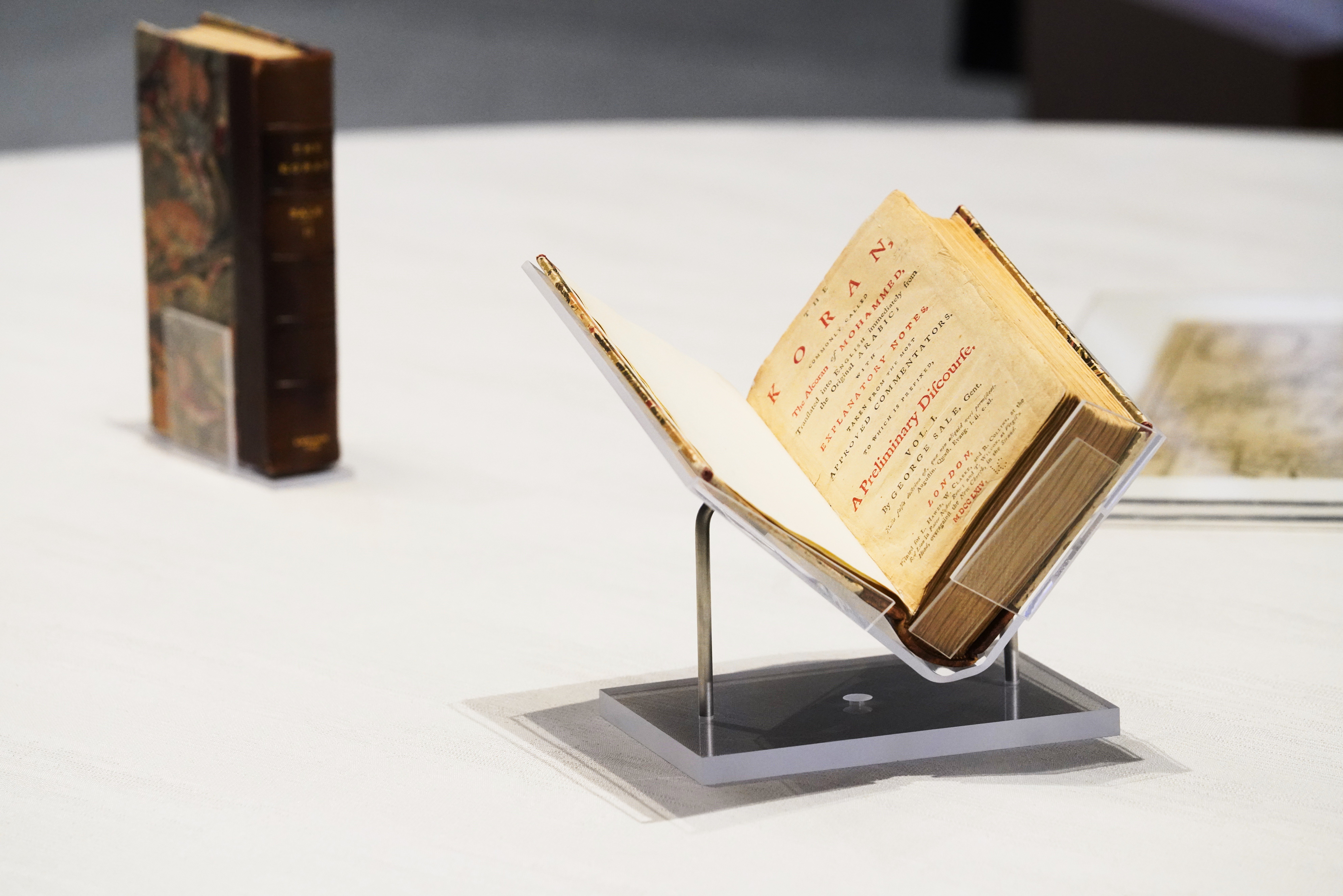 A photo of a display case with two historic books. The book in front is opened, revealing a yellowed first page with the words "Koran" and "A preliminary discourse."