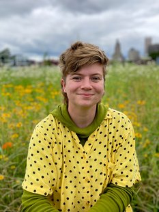 Photograph of MJ Robinson wearing a yellow shirt with black polka dots with a green shirt underneath, sitting in a field of wildflowers.