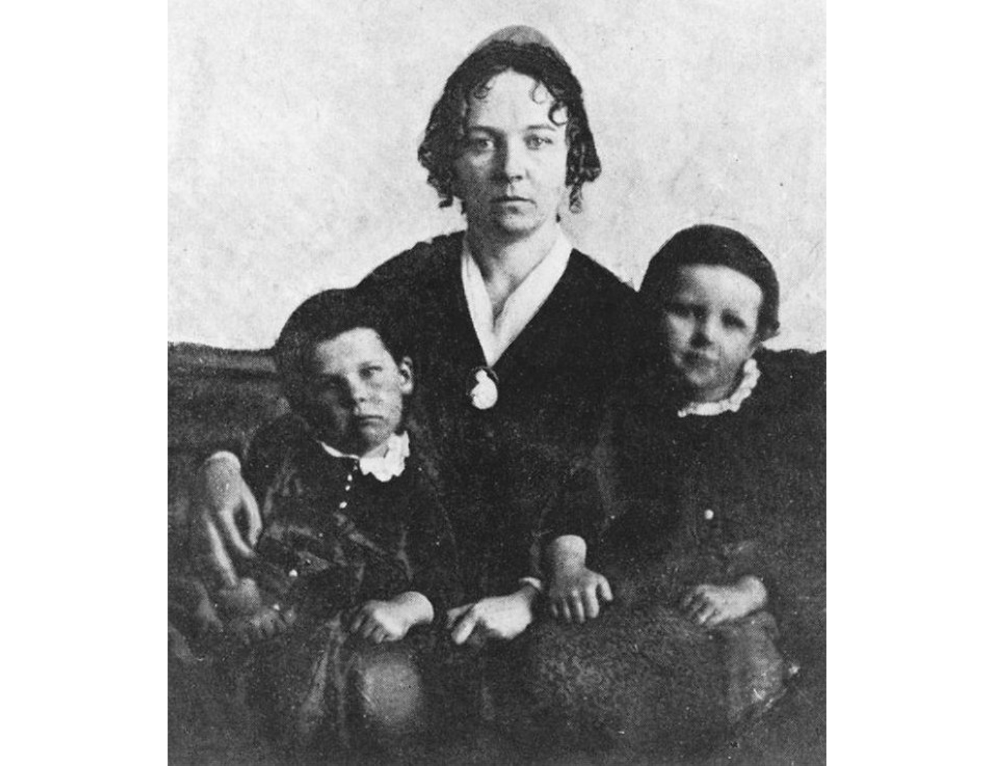 A partially blurry black and white photo of Elizabeth Cady Stanton seated with her two young children on either side. She and one child look seriously into the camera while the other child gives a slight smile.