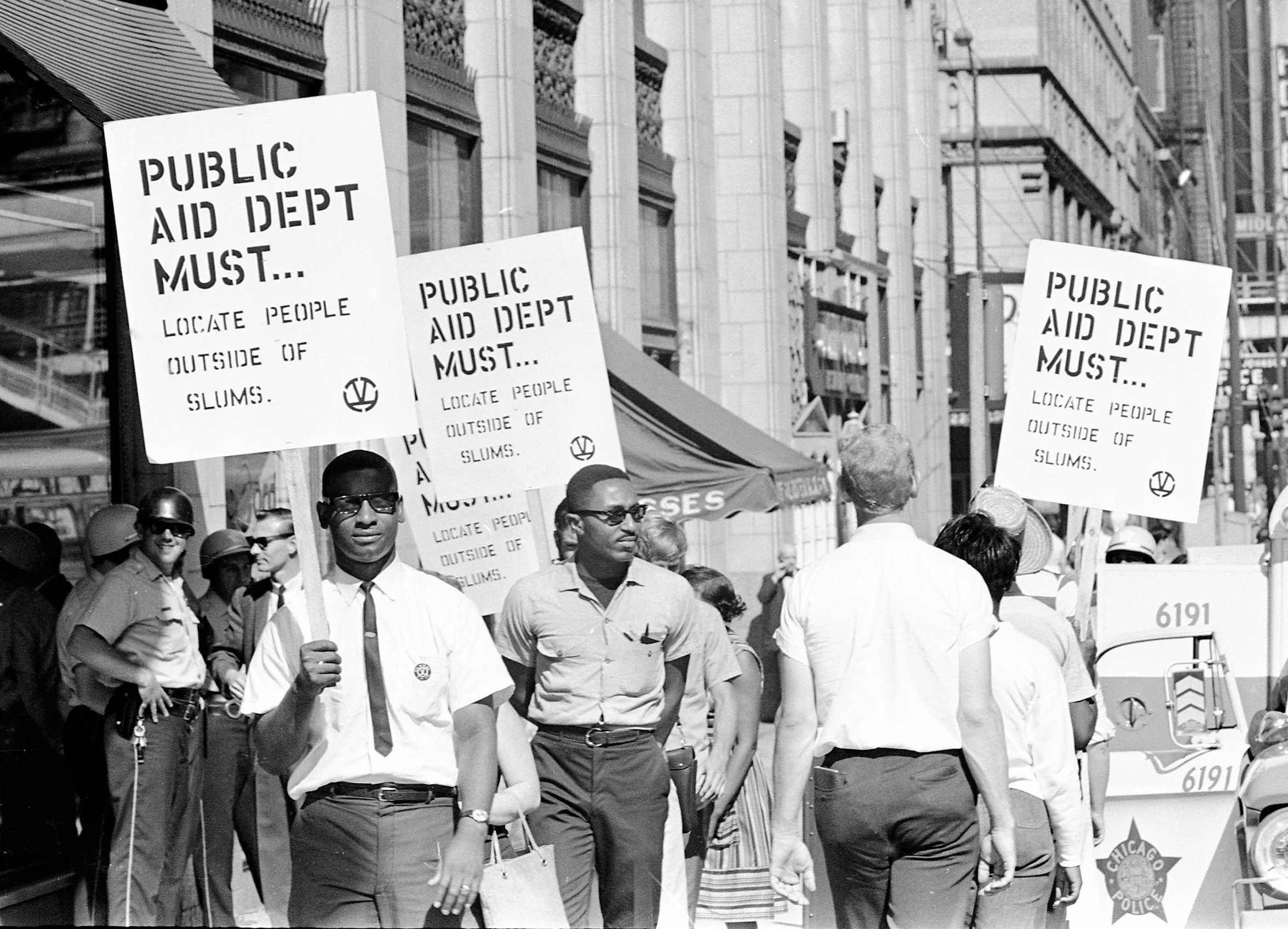 Demonstrators on a crowded sidewalk hold signs that read, "Public Aid Department Must Locate People Outside of Slums."